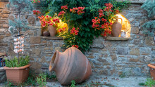 Aegean, Mediterranean Type Garden Consisting Of A Large Old Amphora, Pottery Jugs, Plant In Pot And Trumpet Vine Flowers On The Wall