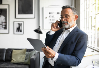 Positive handsome caucasian senior businessman in suit and glasses uses tablet, calls by phone in modern office