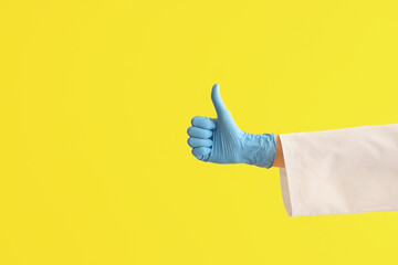 Wall Mural - Female doctor's hand in medical glove showing thumb-up on yellow background