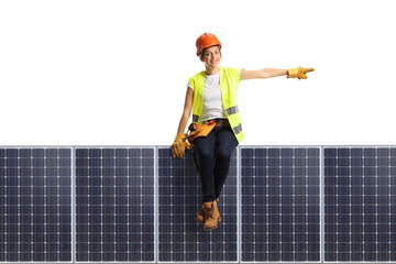 Female construction worker wearing a hardhat and a tool belt sitting on a solar panel and pointing