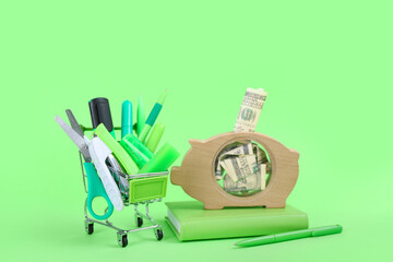 Shopping cart full of different stationery and wooden piggy bank with money on green background