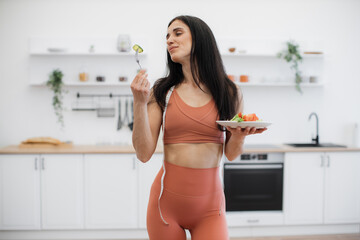 Wall Mural - Portrait of slim beautiful fitness model posing with plate of nutritious snack and smiling at camera. Good-looking woman in tight sport clothing and measuring tape on neck leading healthy lifestyle.