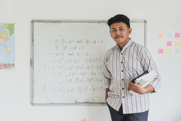Portrait of confident Asian male teacher holding books at the classroom. Knowledge, education and Teachers Day