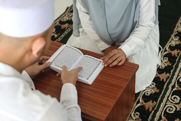 Over shoulder view of Asian muslim man teaching his wife reading Koran or Quran in mosque. Muslim couple reciting Quran together