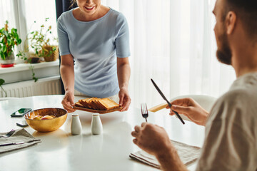 Wall Mural - Smiling woman putting toasts on table near boyfriend with cutlery and smartphones in morning at home