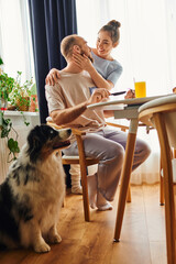 Wall Mural - Smiling woman in homewear hugging boyfriend near breakfast and border collie at home in morning