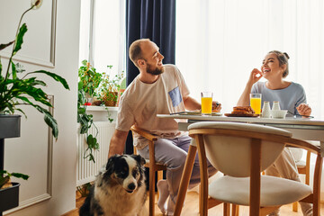 Joyful woman in homewear having tasty breakfast with boyfriend near border collie dog at home