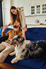 Wall Mural - Positive couple in casual clothes holding hands and spending time near border collie on couch