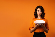 Young Beautiful Stylish Woman Holding An Empty Plate Or Dish Isolated On Orange Background With Copy Space