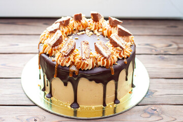 Wall Mural - Peanut butter cream cake with melted chocolate, candy bars, bites of peanuts and salted caramel on wooden background