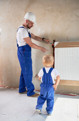 Wall Mural - Male plumber using wrench tool and pointing at radiator while installing heating system together with child. Man and baby boy wearing work overalls while working on home renovation.