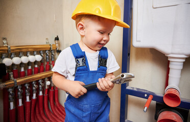 Wall Mural - Baby boy looking at construction tool and smiling while standing near household plumbing installation. Kid in safety helmet and work overalls holding repair instrument wrench. Home renovation concept.