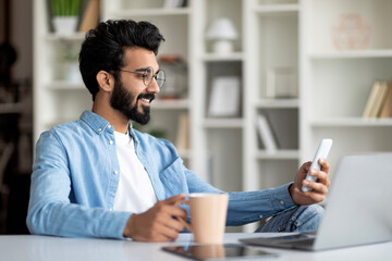 Wall Mural - Indian Male Freelancer Using Smartphone And Drinking Coffee At Home Office