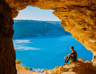 Wall Mural - Gozo Island Malta, a young man and a View of Ramla Bay, from inside Tal Mixta Cave Gozo looking out over the blue ocean on a bright day during winter in Malta