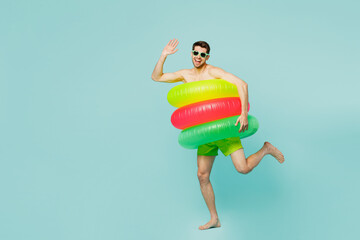 Full body young man wear green shorts swimsuit glasses relax near hotel pool in three inflatable rings look camera wave hand isolated on plain blue background Summer vacation sea rest sun tan concept