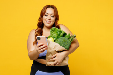 Wall Mural - Young chubby overweight plus size big fat fit woman wear blue top warm up training hold bag with vegetables use mobile cell phone isolated on plain yellow background studio gym. Workout sport concept.