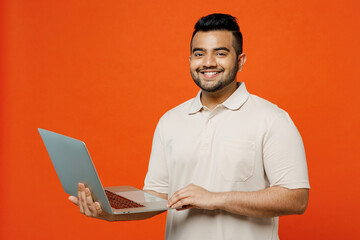 Young fun smiling smart IT Indian man he wears white t-shirt casual clothes hold use work on laptop pc computer look camera isolated on plain orange red background studio portrait. Lifestyle concept.
