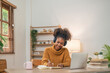 © Natee Meepian - Distance Education. Portrait of smiling woman african american sitting at desk, using laptop and writing in notebook, taking notes, watching tutorial, lecture or webinar, studying online at home