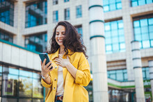 Young Positive Businesswoman With Take Away Coffee Cup In Hand Using Mobile Phone While Standing On Sunny Street Near Office Building, Chatting Online With Friends During Break At Work