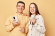 © sementsova321 - Cheerful joyful young man and woman couple standing together isolated over beige background using mobile phone enjoying coffee or tea from thermos.