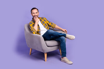 Wall Mural - Photo of positive cheerful man wear trendy clothes relax rest chair hand touch chin enjoy comfort isolated on purple color background