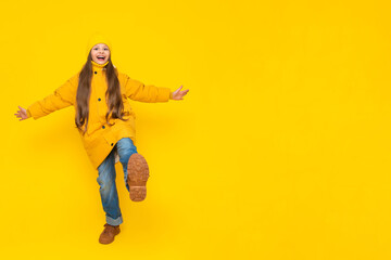 A little girl in full height in an autumn coat, down jackets and hats, rejoices in autumn. The child joyfully raises his leg with his arms outstretched to the sides. Yellow isolated background.