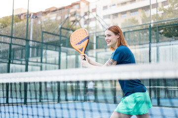 A pretty girl in sportswear is playing a game of outdoor paddle tennis at sunset. The woman is posing with the racket to receive the ball. Concept of women playing paddle.