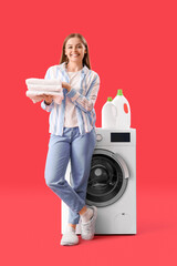 Pretty young woman holding stack of clean towels near washing machine on red background