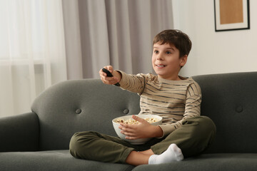 Poster - Little boy holding bowl of popcorn and changing TV channels with remote control on sofa at home