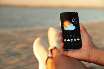 Wall Mural - man checking weather using app on smartphone near river, closeup. data, sun with cloud and other ill