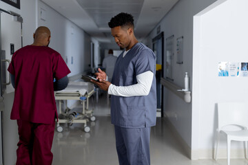 Wall Mural - African american male doctor wearing scrubs using tablet in corridor at hospital