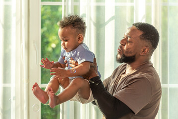 Wall Mural - Happy african black parents dad father throw baby son on piggyback and neck riding teasing fun near window. Black baby son and daddy enjoy teasing kiss cheek and neck riding at window light