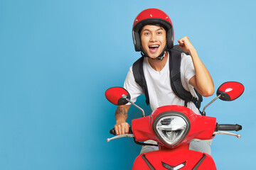 Wall Mural - Asian happy guy in red helmet riding red motorbike and holds one fist up, posing on blue backdrop with wide smile, lifestyle concept, copy space