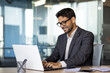 © Liubomir - Young successful Arab businessman in business clothes works inside the office with a laptop, satisfied with the results, the boss in glasses types on the computer keyboard.