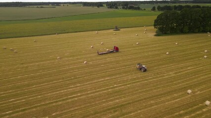 Wall Mural - Aerial footage of a tractor working with cargo truck and hay rolls in a cultivated field with trees