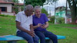 Two diverse seniors looking at phone sitting on park bench outdoors laughing and smiling together. Older friend sharing online media content to companion depicting authentic friendship