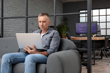 Mature male programmer with modern laptop sitting on sofa in office