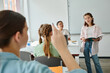 © LIGHTFIELD STUDIOS - Teen schoolgirl holding notebook near classmate raising hand and african american teacher in class