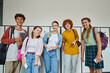 © LIGHTFIELD STUDIOS - cheerful teenage students holding devices and looking at camera in school hallway, friends