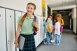 © LIGHTFIELD STUDIOS - cheerful teenage girl holding laptop and looking away in hallway, blur, schoolkids on background