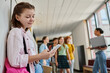 © LIGHTFIELD STUDIOS - happy girl using smartphone, chatting and standing in school hallway, diversity, teacher, kids, blur
