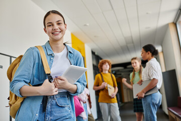 teenage girl with digital tablet looking at camera in school hallway, blurred background, students
