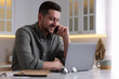 © New Africa - Happy man talking on phone while working with laptop at white table in kitchen