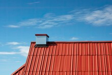A house with a red roof and dormer against a backdrop of clear blue sky. The roof has a decorative metal design and is made from a metal profile. The roofing is covered with stainless steel cladding