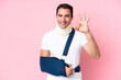 © luismolinero - Young caucasian man with broken arm and wearing a sling isolated on pink background showing ok sign with fingers