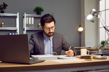 Busy young man who works as auditor, bookkeeper or financial accountant in company is sitting at office desk, doing paperwork, reading business documents, holding pen, taking notes in accounting book