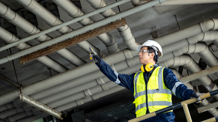 Asian male building inspection engineer pointing at plumber piping system. Construction worker man with reflective vest, safety helmet, protective goggles and ear muffs working at at construction site