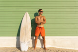 Fototapeta Natura - A smiling man with nice body wearing sunglasses standing at the light green with a surfboard - looking to the side