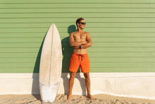 A Smiling Man With Nice Body Wearing Sunglasses Standing At The Light Green With A Surfboard - Looking To The Side