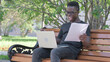 © stockbakers - Young African Man Celebrating while Working on Laptop and Documents Outdoor